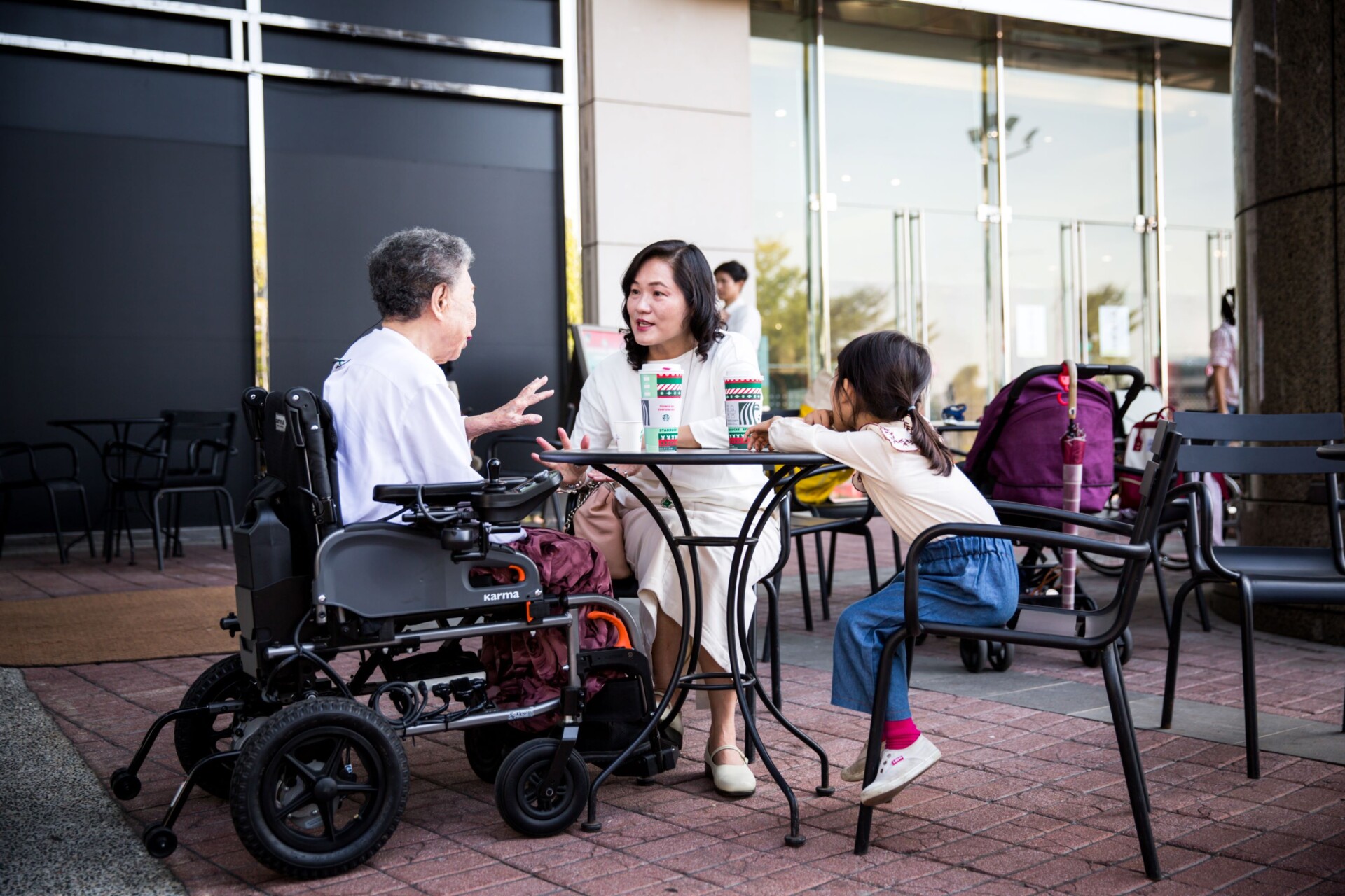 An-elder-in-a-travel-power-wheelchair-sitting-outdoor-and-having-coffee-with-her-family