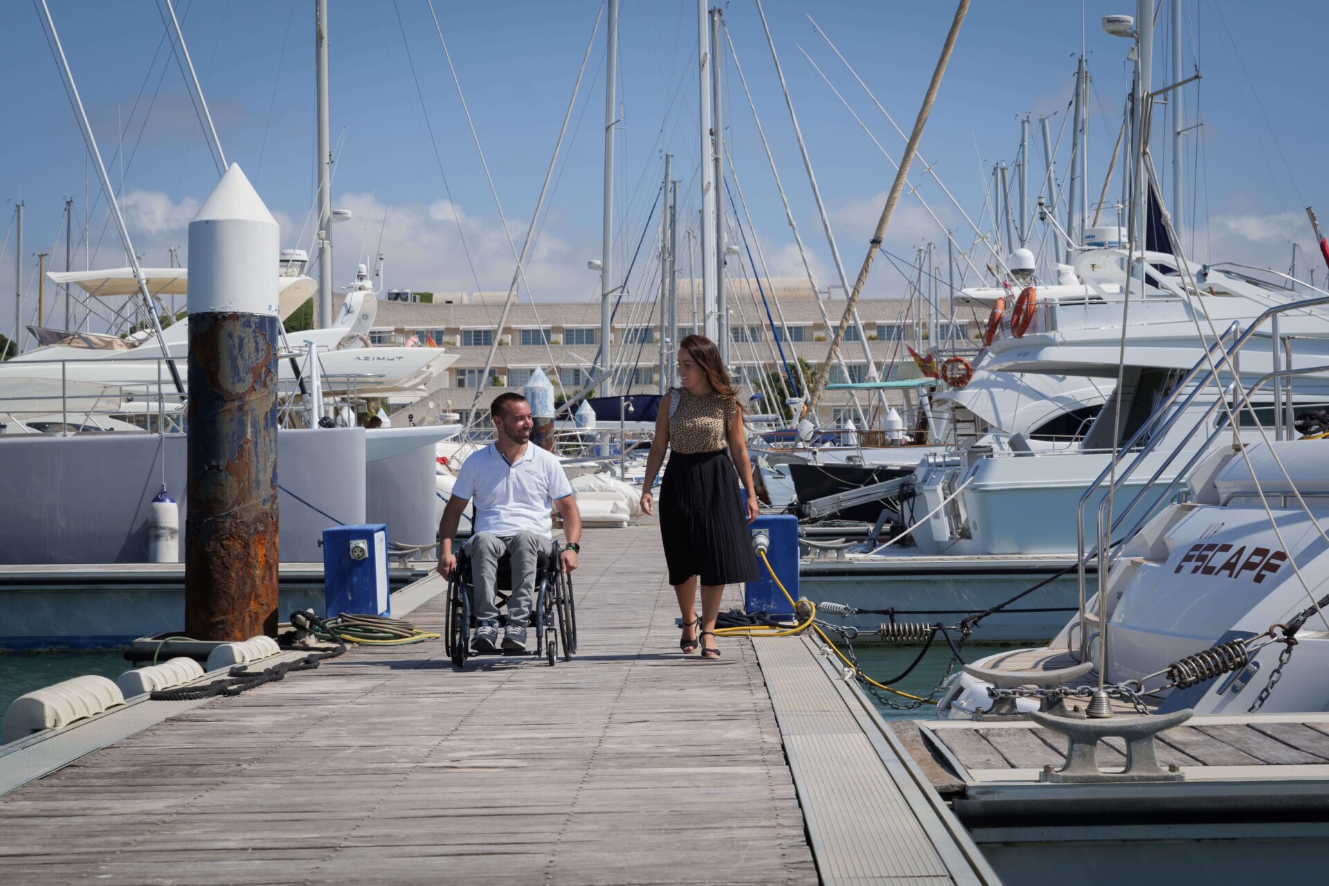 A man in a wheelchair and a woman walking on a marina dock surrounded by yachts on a sunny day